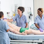 Two students and one lecturer all dressed in nurse skills uniform are standing around a mannequin on a bed