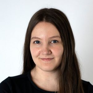 Student Erin smiles in front of a white backdrop, wearing a black tee. She wears minimal make up and her long hair is half tucked behind her shoulder.