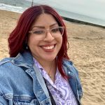 Zoey smiles for a selfie wearing a denim jacket, her red hair is curled, she's located at Bournemouth beach - the sea is in the background.