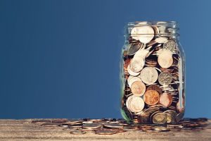 A full jar of coin money with coins spread around the bottom of the jar 