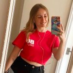Katy stands in front of a mirror holding up her phone taking a selfie wearing a BU branded red top