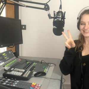 Smiling girl, sitting, holding up her fingers in peace signs, in the Nerve Radio station. There is a mic and radio desk in front of her.