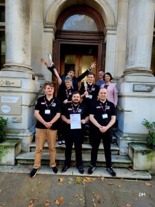 Group of people standing outside a building arch with the person in the front holding a certificate 